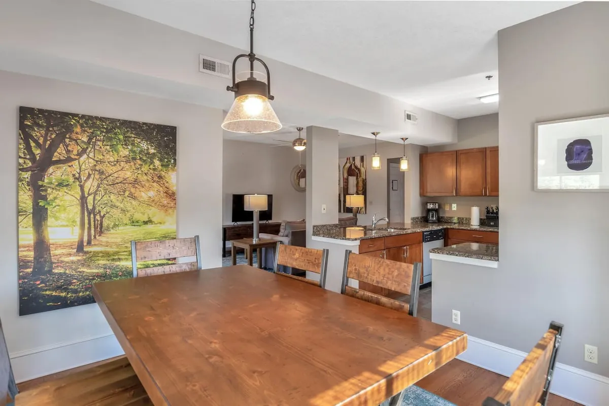 A spacious dining room with a large wooden table, pendant lights, and art depicting a tree-lined path. The setting hints at lively meals and memorable moments shared around the table, blending a sense of home with anticipation for culinary exploration.