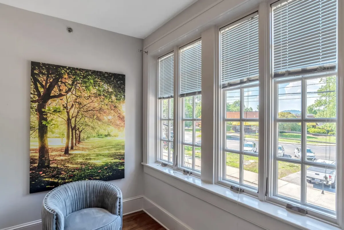 This serene scene features a comfortable chair situated near bright windows, with a large artwork of a tree-lined path on display. The room is filled with natural light, creating an inviting atmosphere for relaxation and reflection, while seasonal changes can be appreciated through the windows.