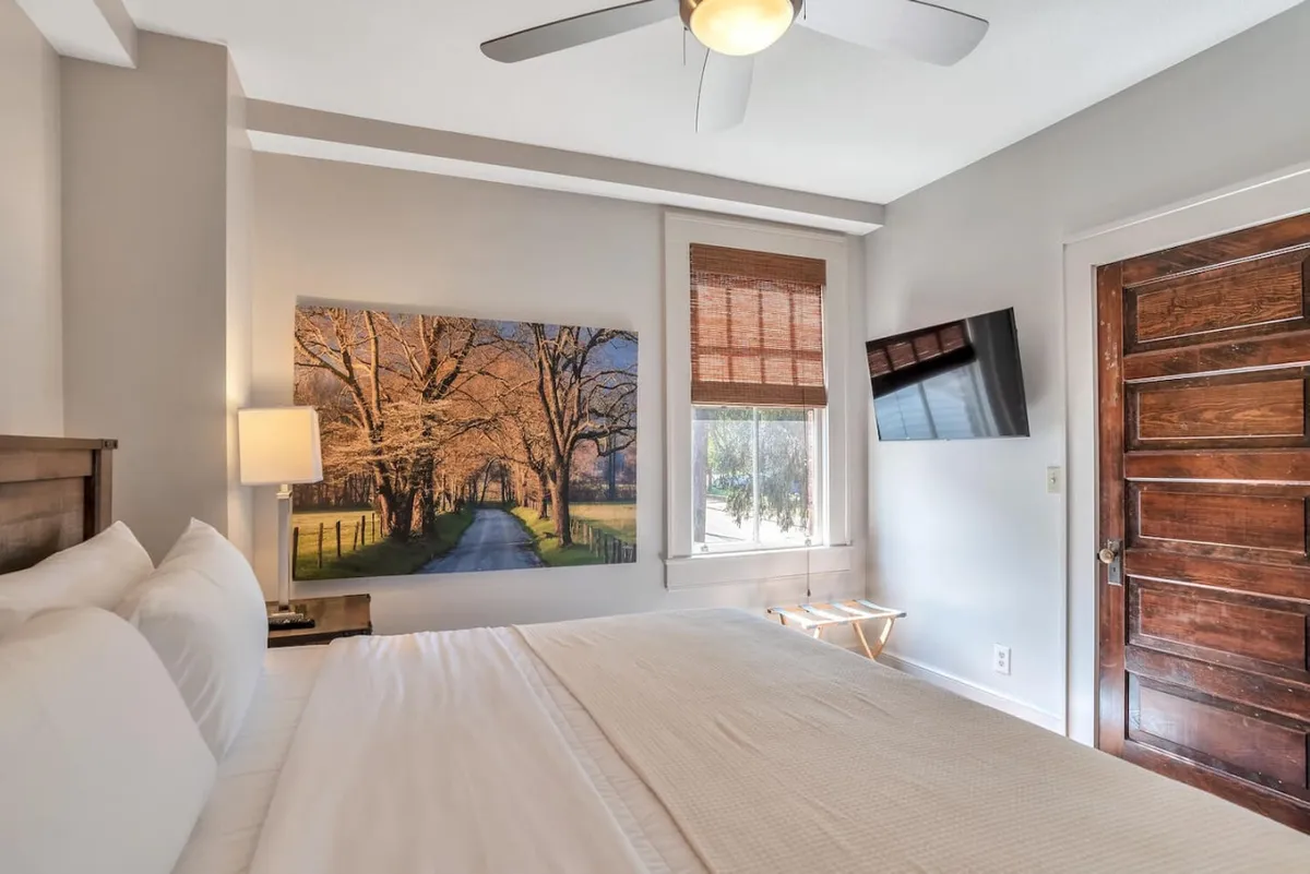 A cozy bedroom featuring a comfortable bed with crisp white linens, a large artwork of a tree-lined path above the headboard, and a classic wooden door. Sunlight pours into the room, emphasizing the peaceful ambiance and the inviting space for relaxation or reading.