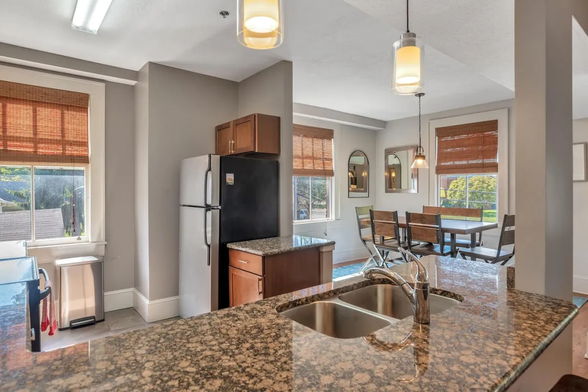 The image features a spacious kitchen with a granite countertop, kitchen island, dining table, and large windows. The setup suggests a seamless transition from meal preparation to dining and socializing, with an abundance of natural light.