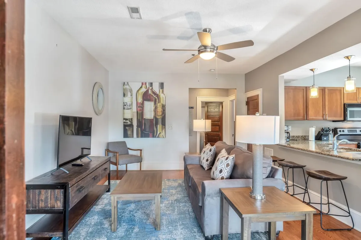 This image showcases a comfortable open living area with a plush sofa decorated with colorful cushions, a large painting of wine bottles that adds character and warmth, a coffee table, television, and bar stools. The scene suggests a relaxed, social vibe perfect for unwinding or entertaining after a day in the city.