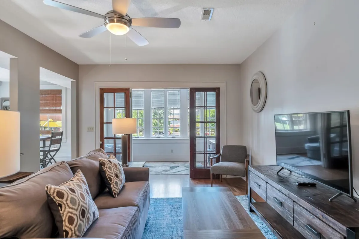 The image shows a cozy living area with a stylish sofa adorned with patterned cushions, a warm lamp casting soft light, a flat-screen television perfect for movie nights, and a glimpse into the inviting dining area beyond. Sunlight gently fills the space, creating a cheerful and welcoming ambiance ideal for relaxation or intimate meals.