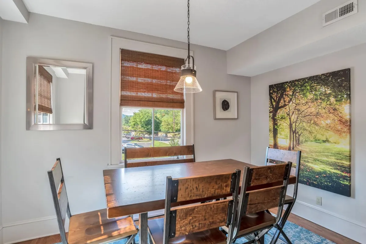 A cozy dining area featuring a wooden table surrounded by stylish chairs, with a large window letting in morning light. Framed artwork and a mirror adorn the walls, suggesting a welcoming space for meals or gatherings.