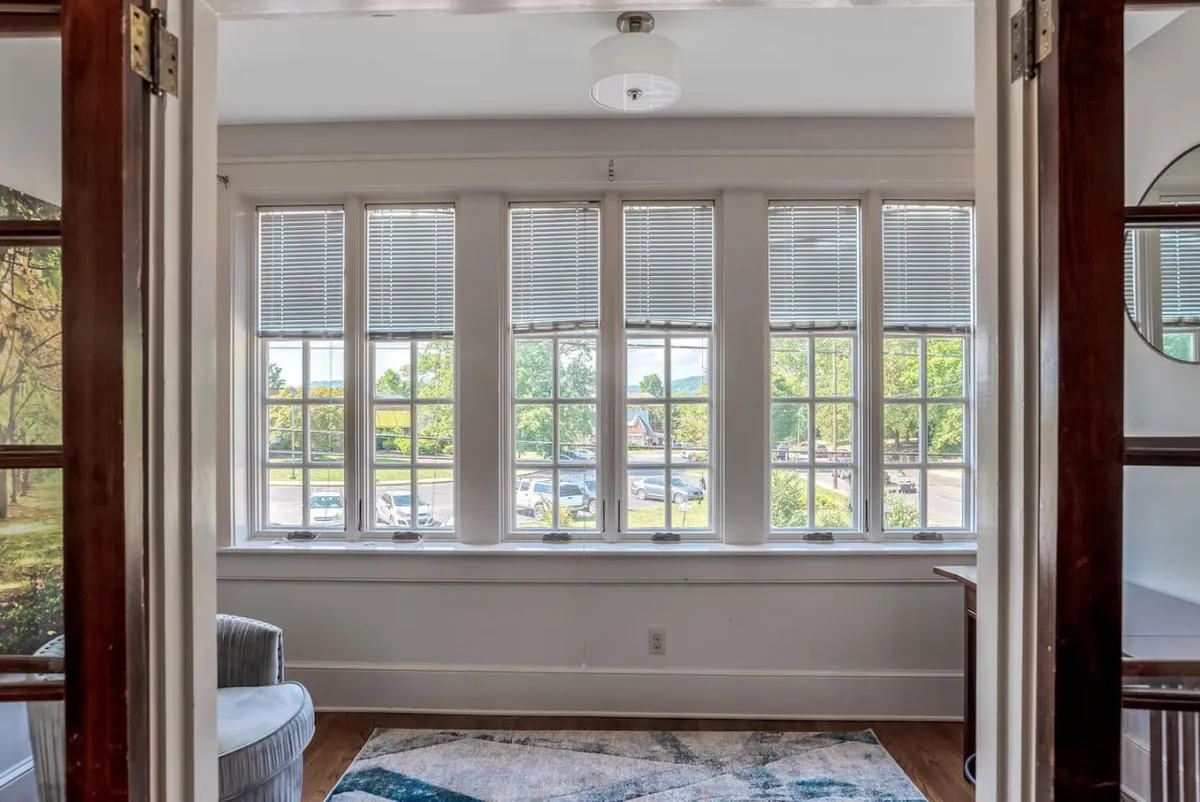 This image features large windows that flood the room with natural light, a colorful rug, and a comfortable chair situated perfectly for enjoying downtown views. The space feels bright and welcoming, with the city just beyond the glass—a nod to the lively urban energy and mentioned construction activity.