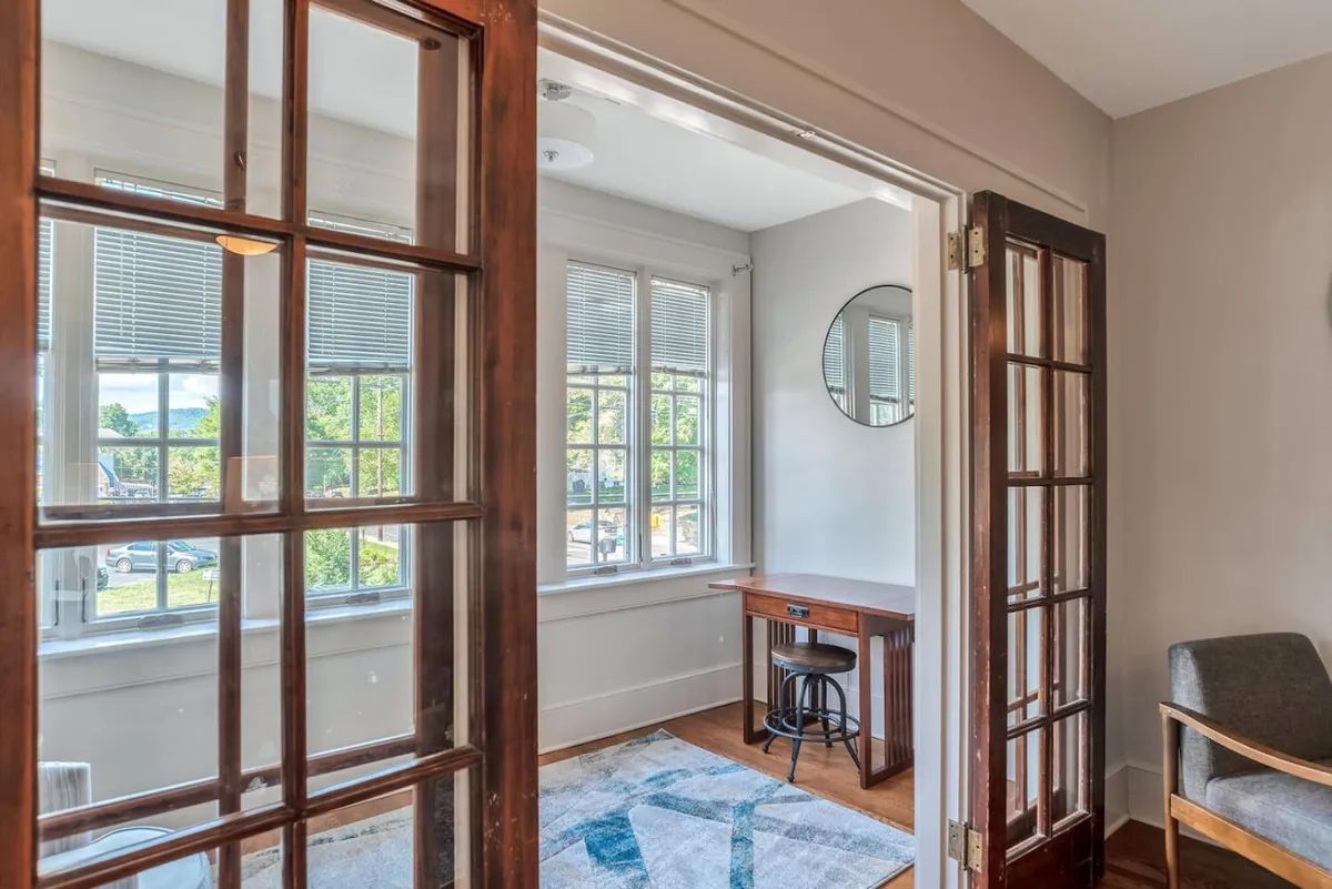 This image features an interior entry space with prominent wooden doors, large windows allowing natural light, a desk, a round mirror, and a patterned area rug. The scene is oriented toward the entrance and exudes a sense of arrival and organization.